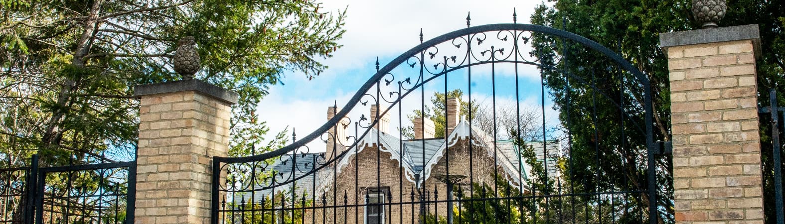 Elegant black wrought iron driveway gates with decorative scrollwork and arched tops, mounted between brick gate pillars to create a secure and stylish entrance to a traditional residential property.